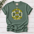 A sage green short-sleeve t-shirt with a vintage-style graphic reading 'FUELED BY PICKLES' around a pickle jar illustration. The shirt is laid flat on a white wood plank background with a gold necklace draped over the shoulders.
