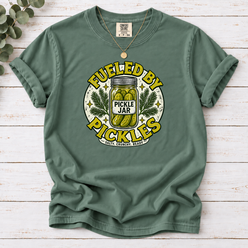 A sage green short-sleeve t-shirt with a vintage-style graphic reading 'FUELED BY PICKLES' around a pickle jar illustration. The shirt is laid flat on a white wood plank background with a gold necklace draped over the shoulders.