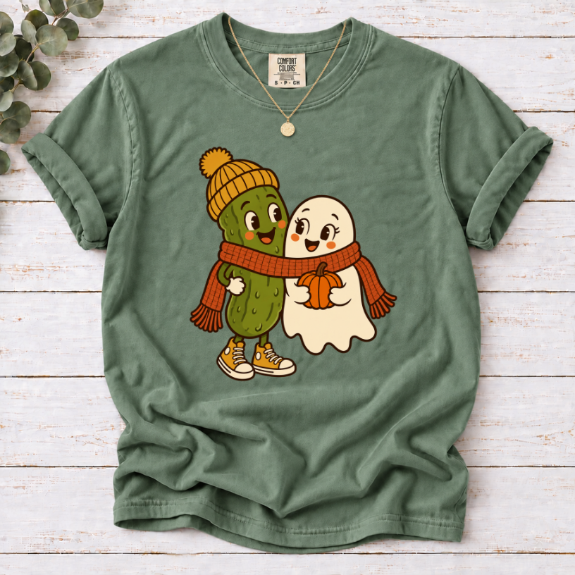 moss T-shirt featuring a smiling pickle in a yellow beanie beside a ghost holding a pumpkin, sharing an orange scarf.”