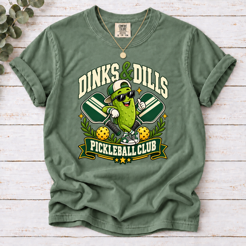 Moss Dinks & Dills Pickleball Club graphic T-shirt featuring a smiling pickle mascot wearing sunglasses and a backward cap, crossed pickleball paddles, and retro collegiate lettering.