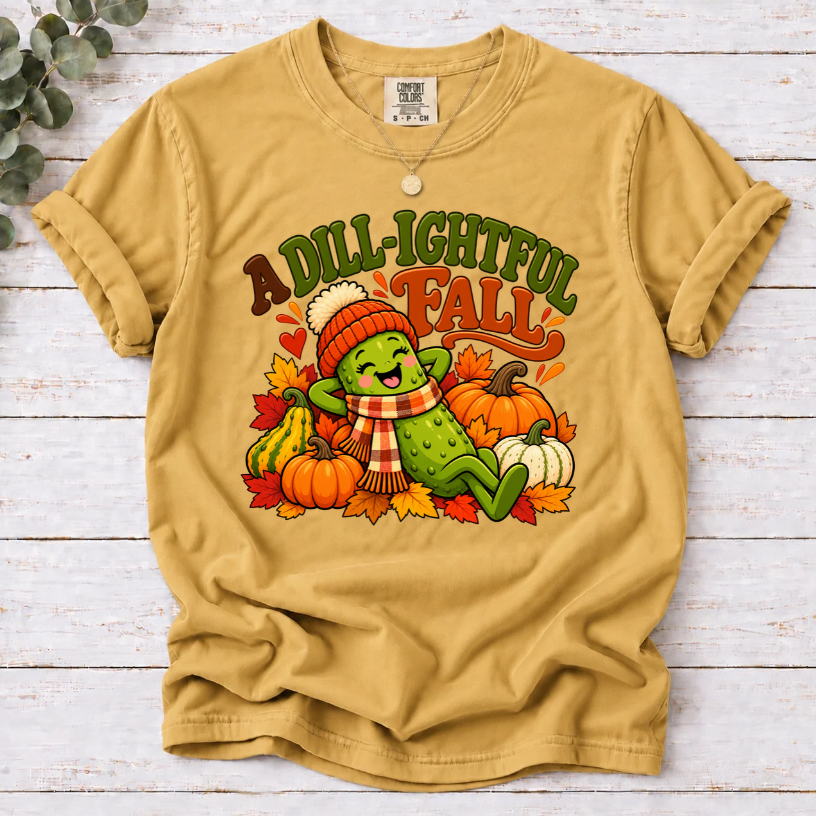 Mustard A Dill-ightful Fall T-shirt with a smiling pickle in a beanie and plaid scarf among pumpkins and autumn leaves.