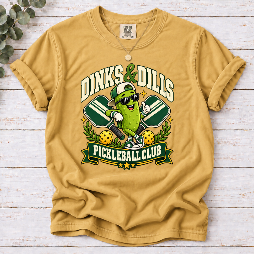 Mustard Dinks & Dills Pickleball Club graphic T-shirt featuring a smiling pickle mascot wearing sunglasses and a backward cap, crossed pickleball paddles, and retro collegiate lettering.