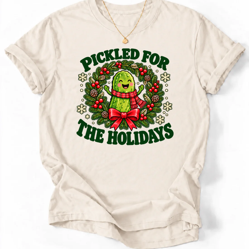 Natural Pickled for the Holidays’ T-shirt with a smiling pickle in a red scarf inside a Christmas wreath.”