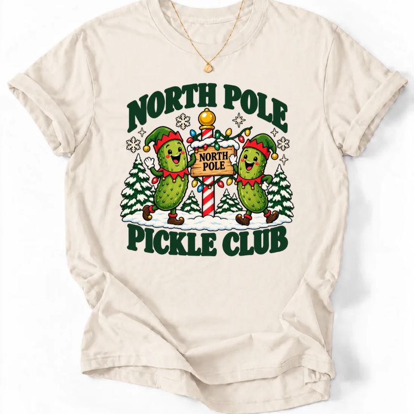 Natural T-shirt with “North Pole Pickle Club” lettering and two pickle elves beside a candy cane signpost and snowy trees.