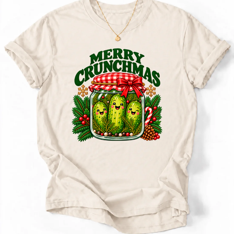 Natural T-shirt with “Merry Crunchmas” lettering above three smiling pickles in a jar decorated with a red bow and Christmas greenery.