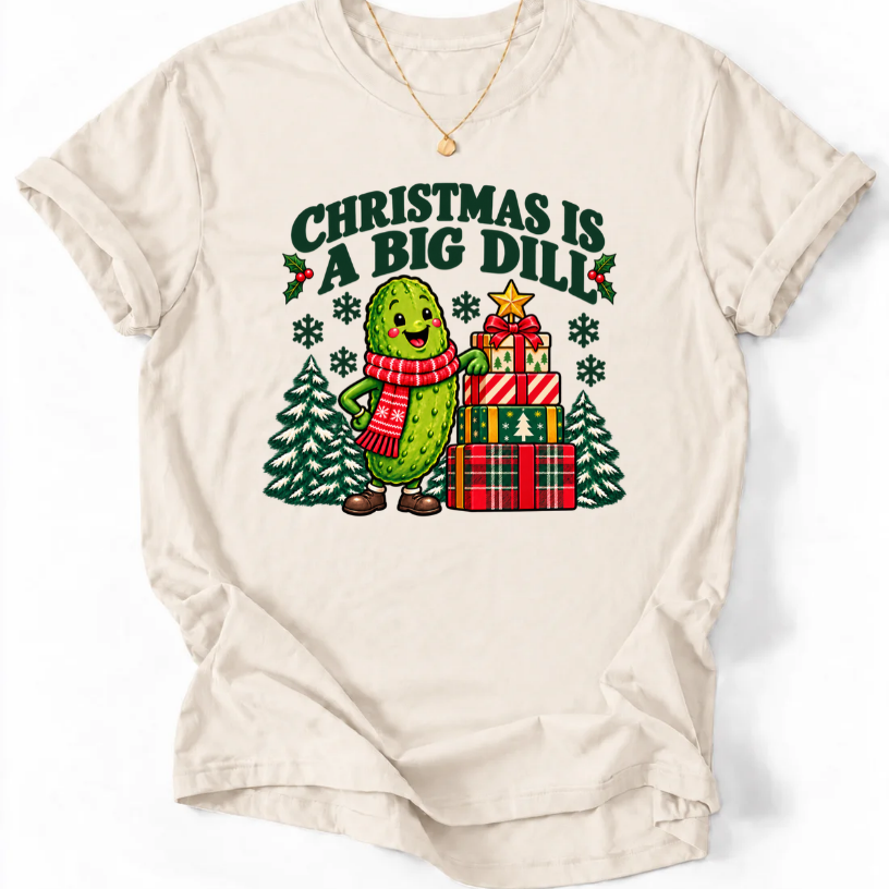 Natural T-shirt with “Christmas Is a Big Dill” lettering and a smiling pickle in a red scarf beside stacked gifts and snowy trees.