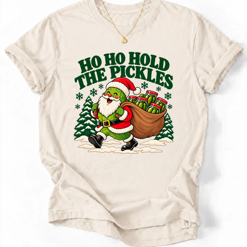 Natural T-shirt with “Ho Ho Hold the Pickles” lettering and a pickle dressed as Santa carrying a sack of pickle jars beside snowy trees.