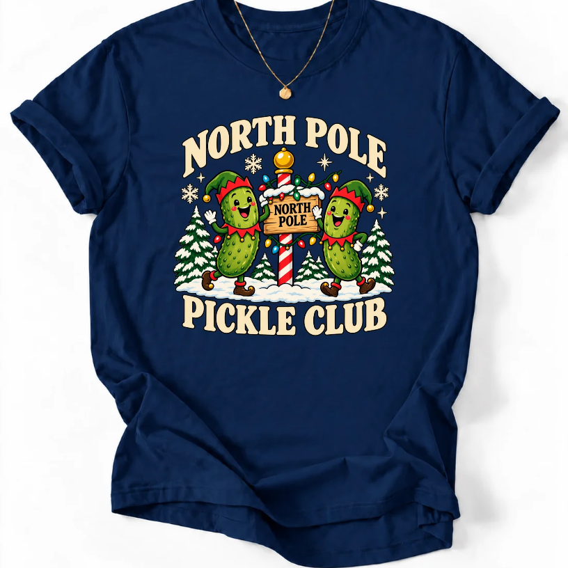 Navy T-shirt with “North Pole Pickle Club” lettering and two pickle elves beside a candy cane signpost and snowy trees.