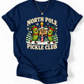 Navy T-shirt with “North Pole Pickle Club” lettering and two pickle elves beside a candy cane signpost and snowy trees.