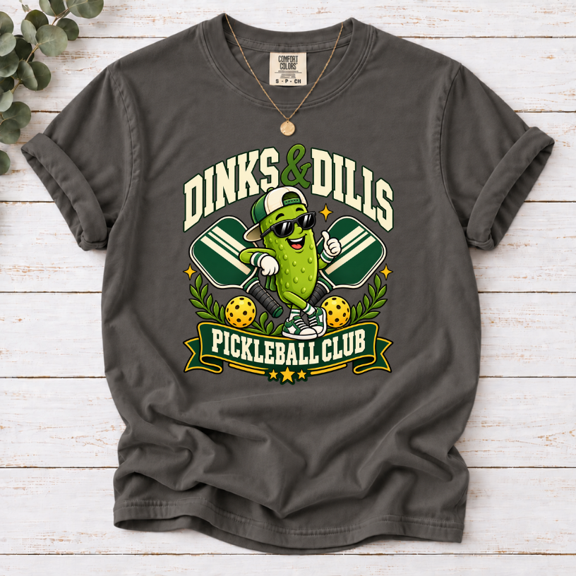 Pepper Dinks & Dills Pickleball Club graphic T-shirt featuring a smiling pickle mascot wearing sunglasses and a backward cap, crossed pickleball paddles, and retro collegiate lettering.