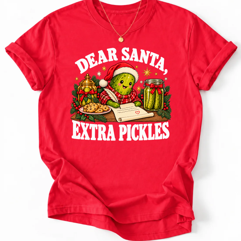 Red ‘Dear Santa, Extra Pickles’ T-shirt featuring a Santa-hat pickle writing a letter beside cookies and pickles.”