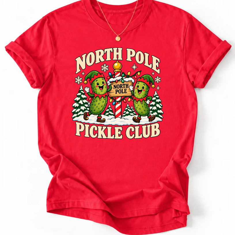 Red T-shirt with “North Pole Pickle Club” lettering and two pickle elves beside a candy cane signpost and snowy trees.