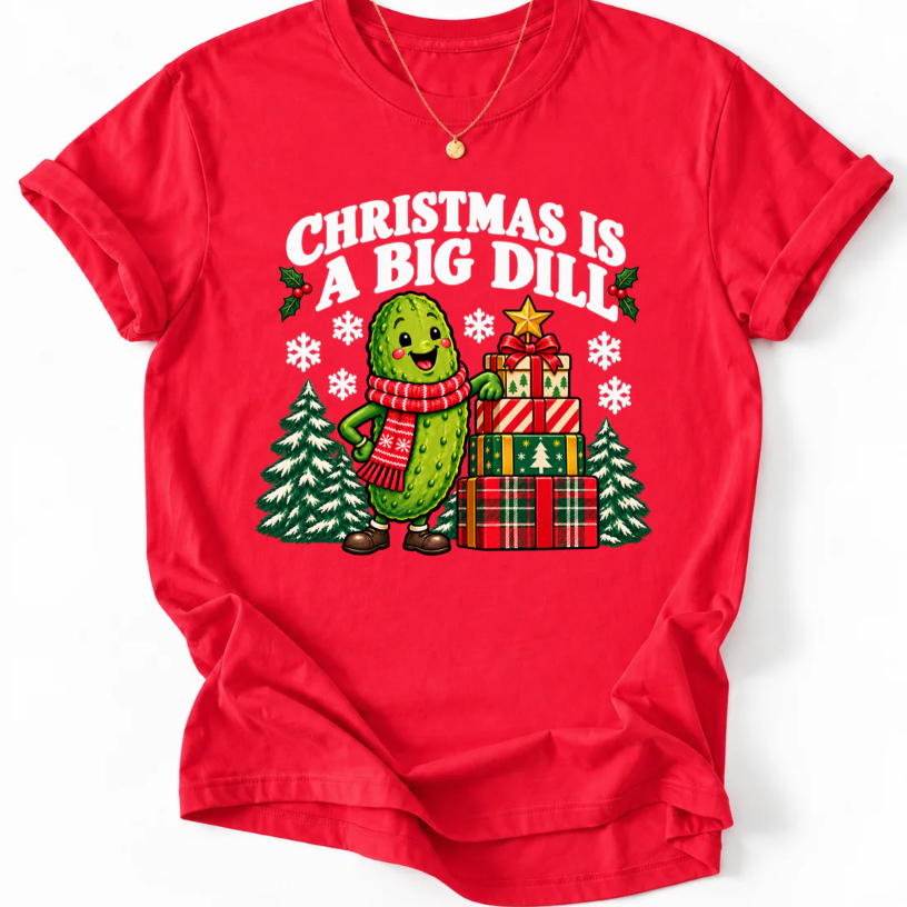 Red T-shirt with “Christmas Is a Big Dill” lettering and a smiling pickle in a red scarf beside stacked gifts and snowy trees.