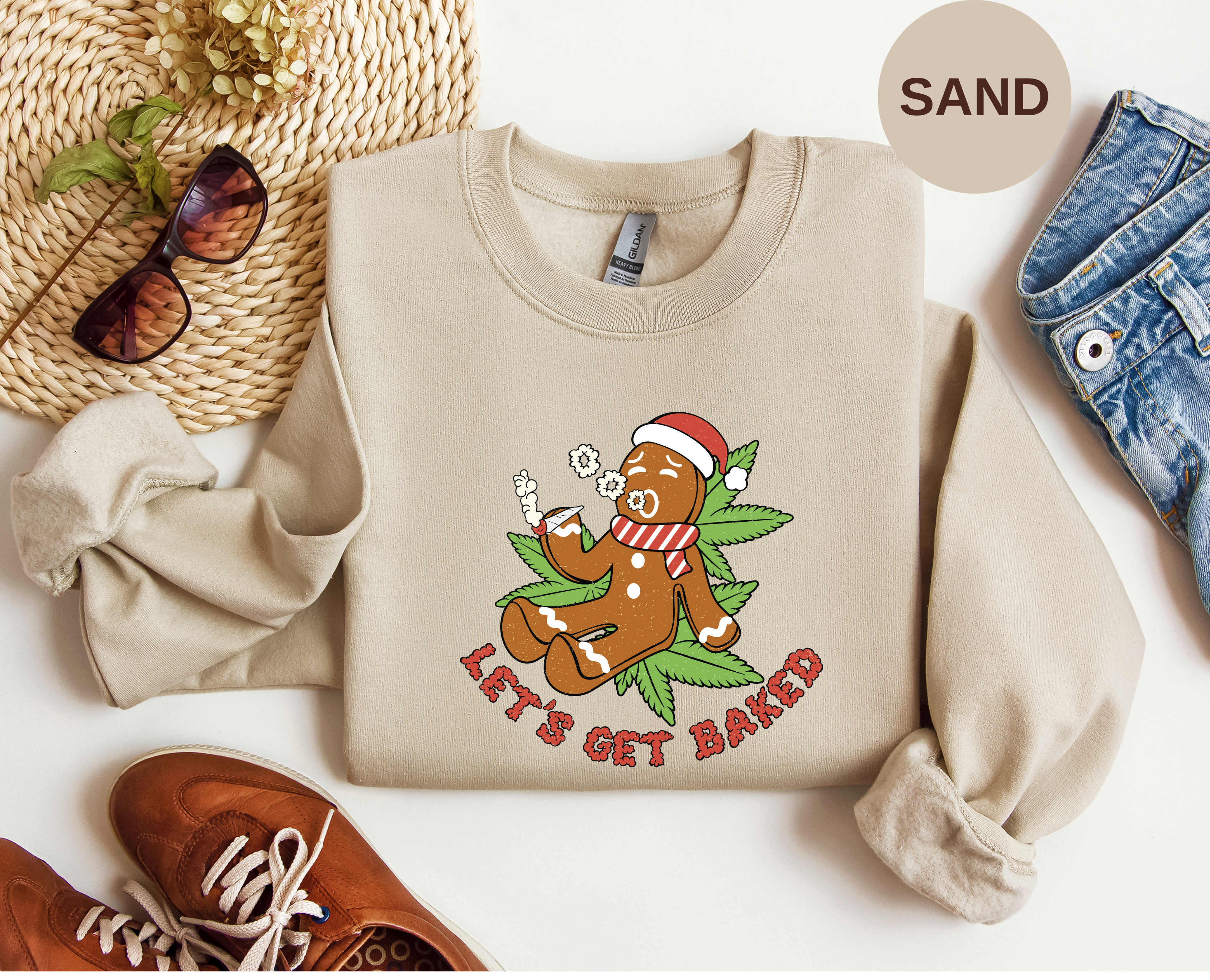A flat-lay of a sand-colored crewneck sweatshirt featuring a gingerbread man graphic with the text 'LET'S GET BAKED' and marijuana The sweatshirt is styled with brown shoes, a woven hat, sunglasses, and blue jeans on a white background, with a 'SAND' color circle overlay.