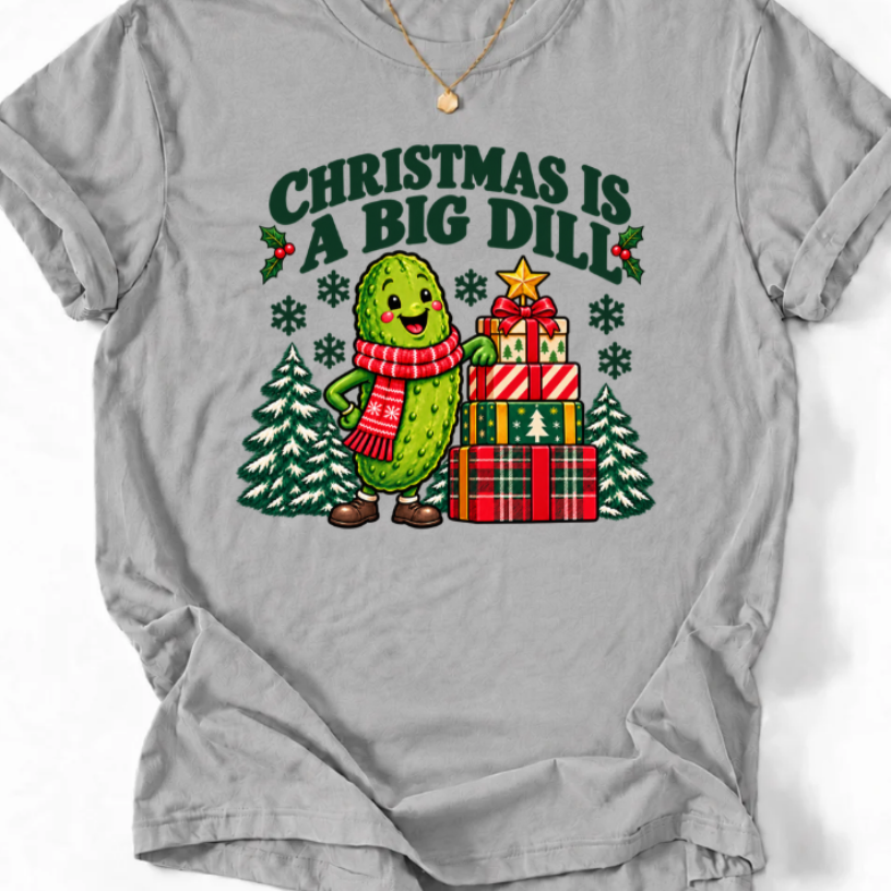 Sport Grey T-shirt with “Christmas Is a Big Dill” lettering and a smiling pickle in a red scarf beside stacked gifts and snowy trees.