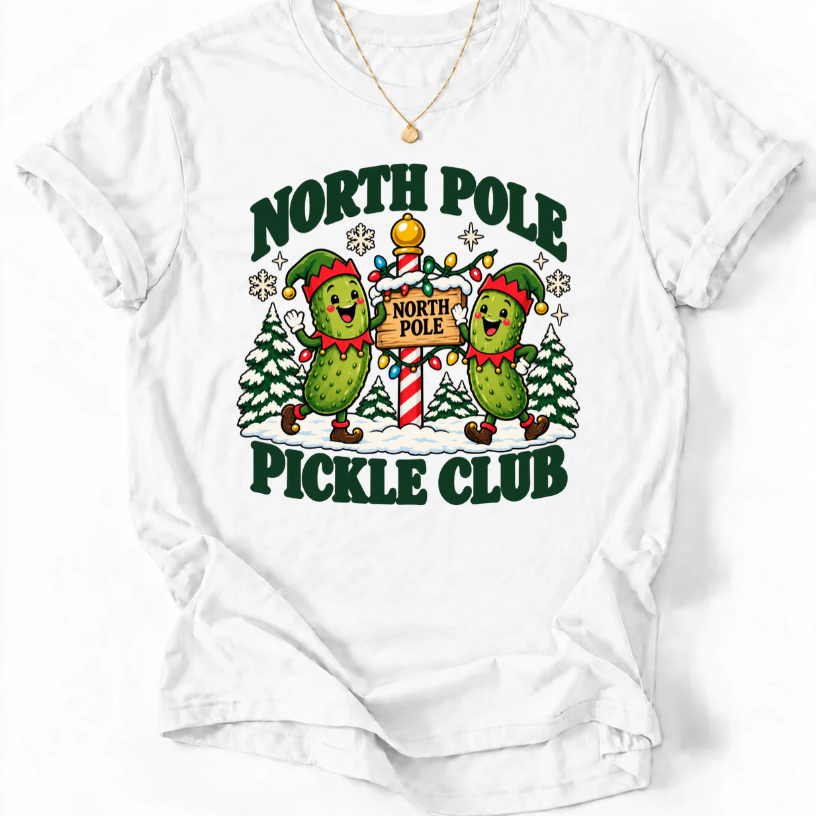 White T-shirt with “North Pole Pickle Club” lettering and two pickle elves beside a candy cane signpost and snowy trees.