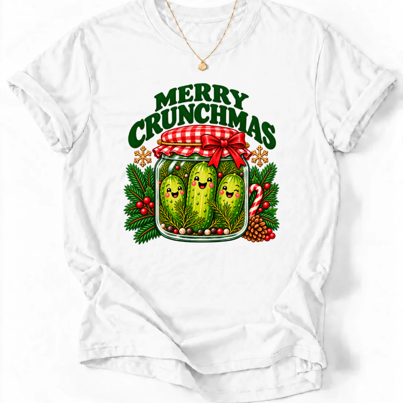 White T-shirt with “Merry Crunchmas” lettering above three smiling pickles in a jar decorated with a red bow and Christmas greenery.
