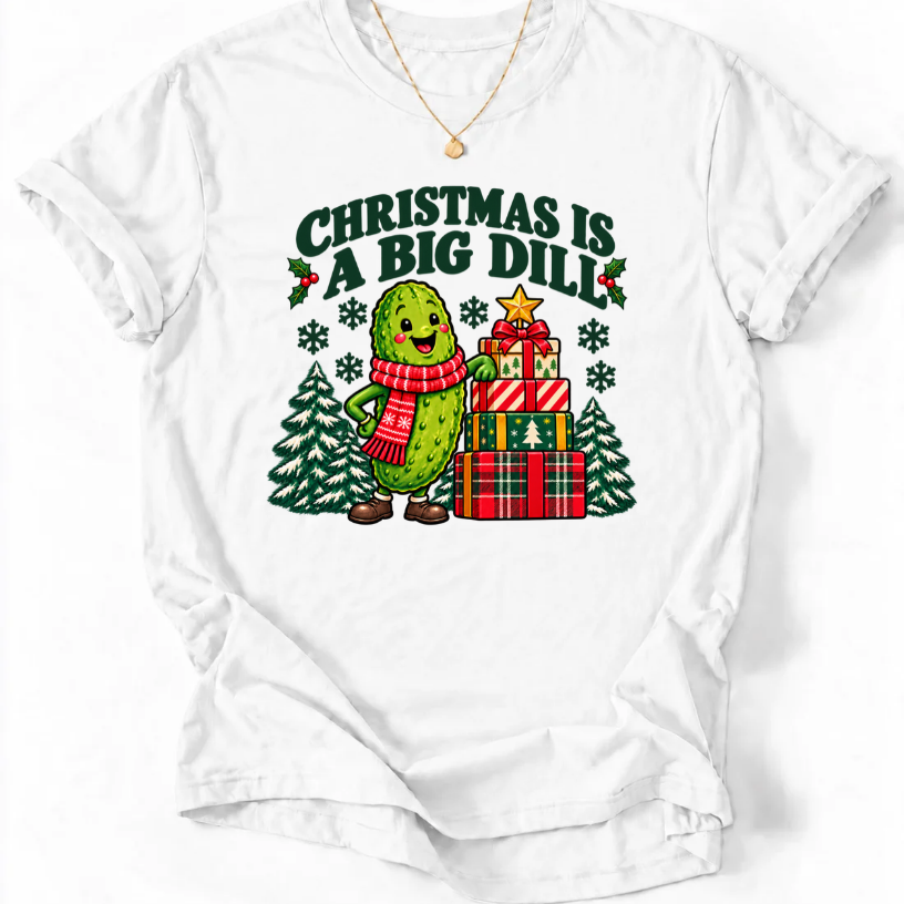White T-shirt with “Christmas Is a Big Dill” lettering and a smiling pickle in a red scarf beside stacked gifts and snowy trees.