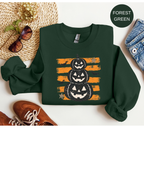 A dark forest green long-sleeve sweatshirt laid flat on a white background, featuring a graphic of three stacked jack-o'-lantern pumpkins over orange distressed stripes. Styled flat-lay props include brown shoes, a woven hat with sunglasses, blue jeans, and a small circular label reading FOREST GREEN.