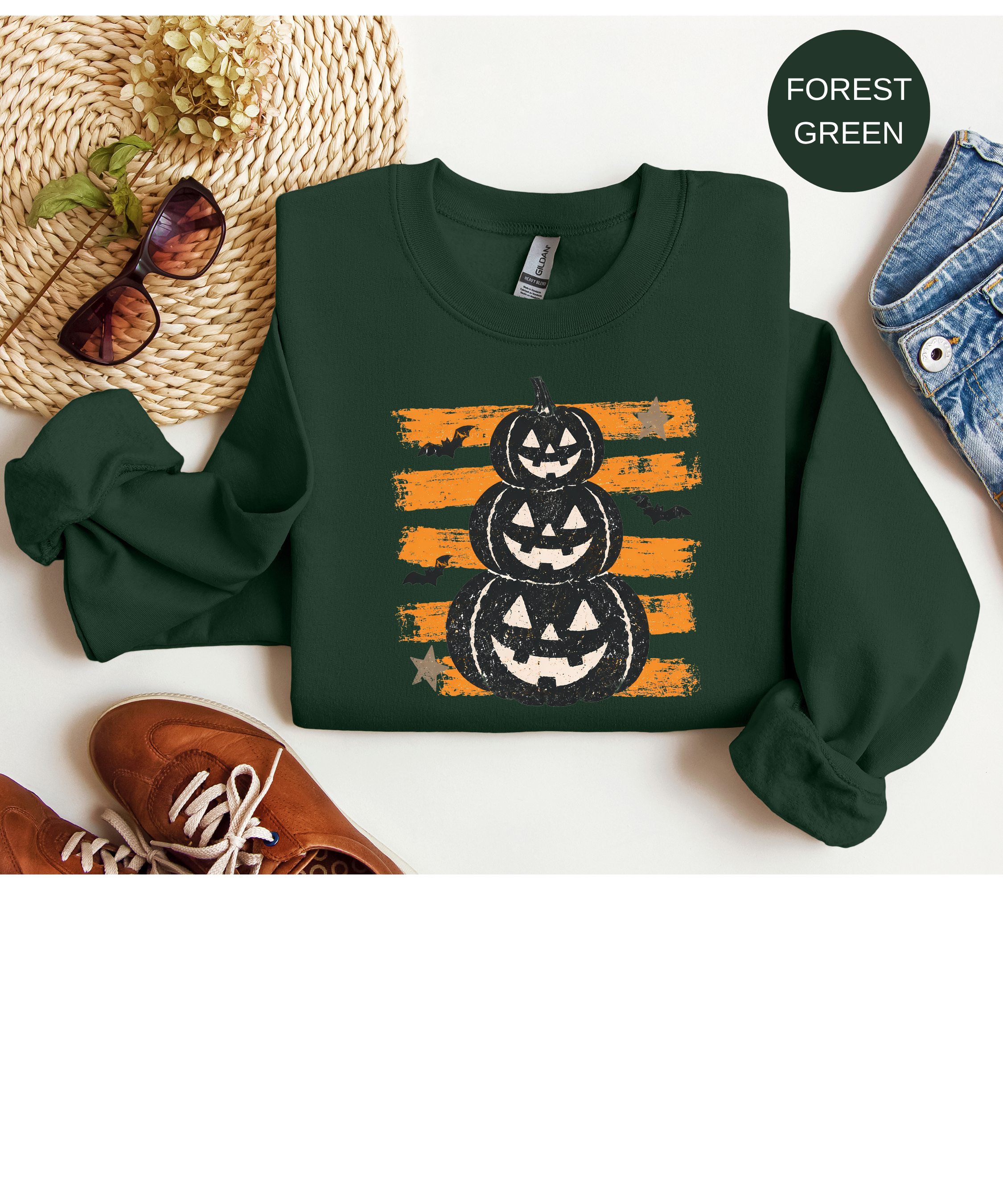 A dark forest green long-sleeve sweatshirt laid flat on a white background, featuring a graphic of three stacked jack-o'-lantern pumpkins over orange distressed stripes. Styled flat-lay props include brown shoes, a woven hat with sunglasses, blue jeans, and a small circular label reading FOREST GREEN.