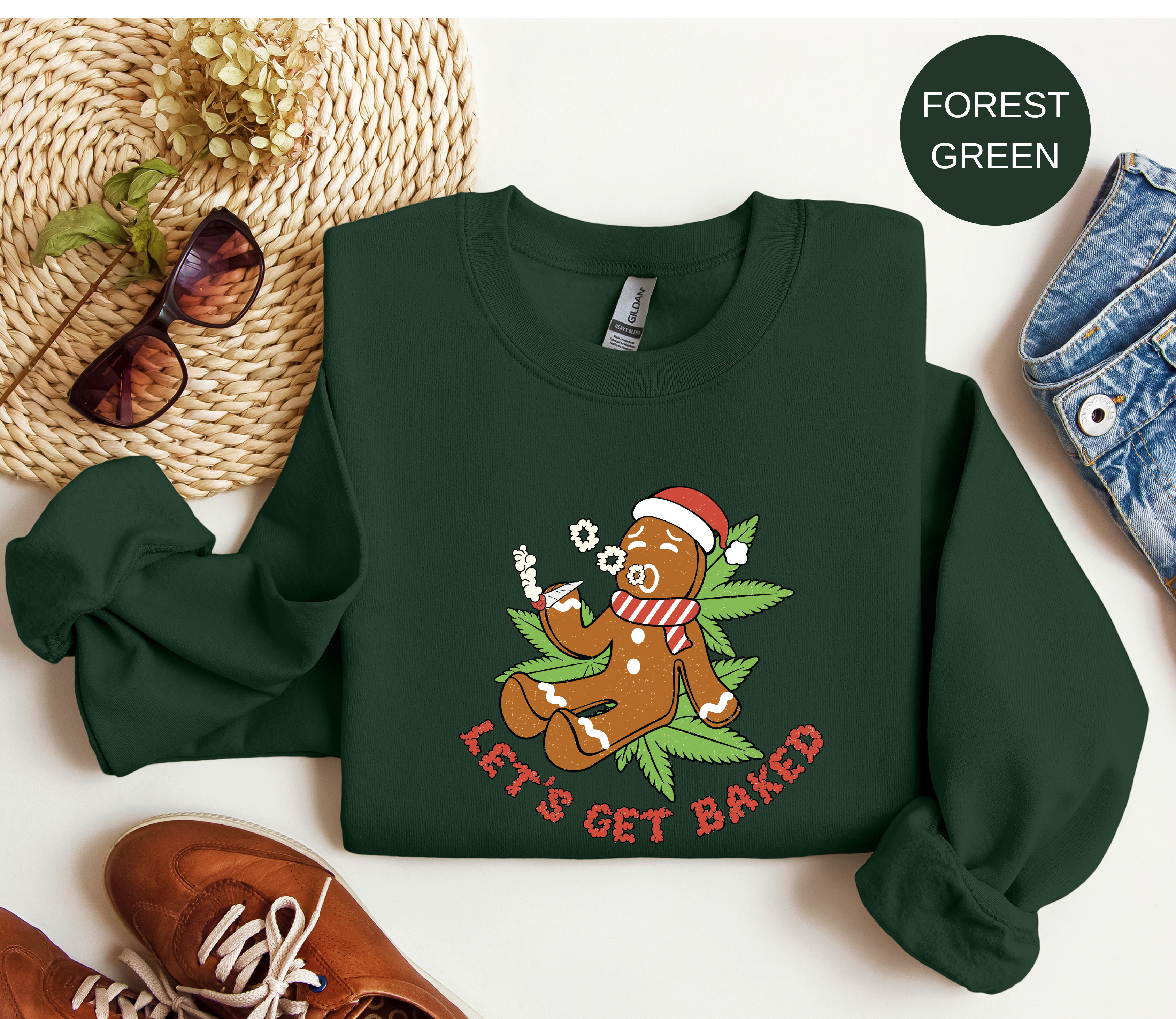 A dark forest green crewneck sweatshirt laid flat on a white background, styled with a straw hat, sunglasses, brown shoes, and blue jeans. The sweatshirt features a graphic of a gingerbread man wearing a Santa hat and scarf, sitting on green leaves and smoking a spliff with the text 'LET'S GET BAKED' in red.