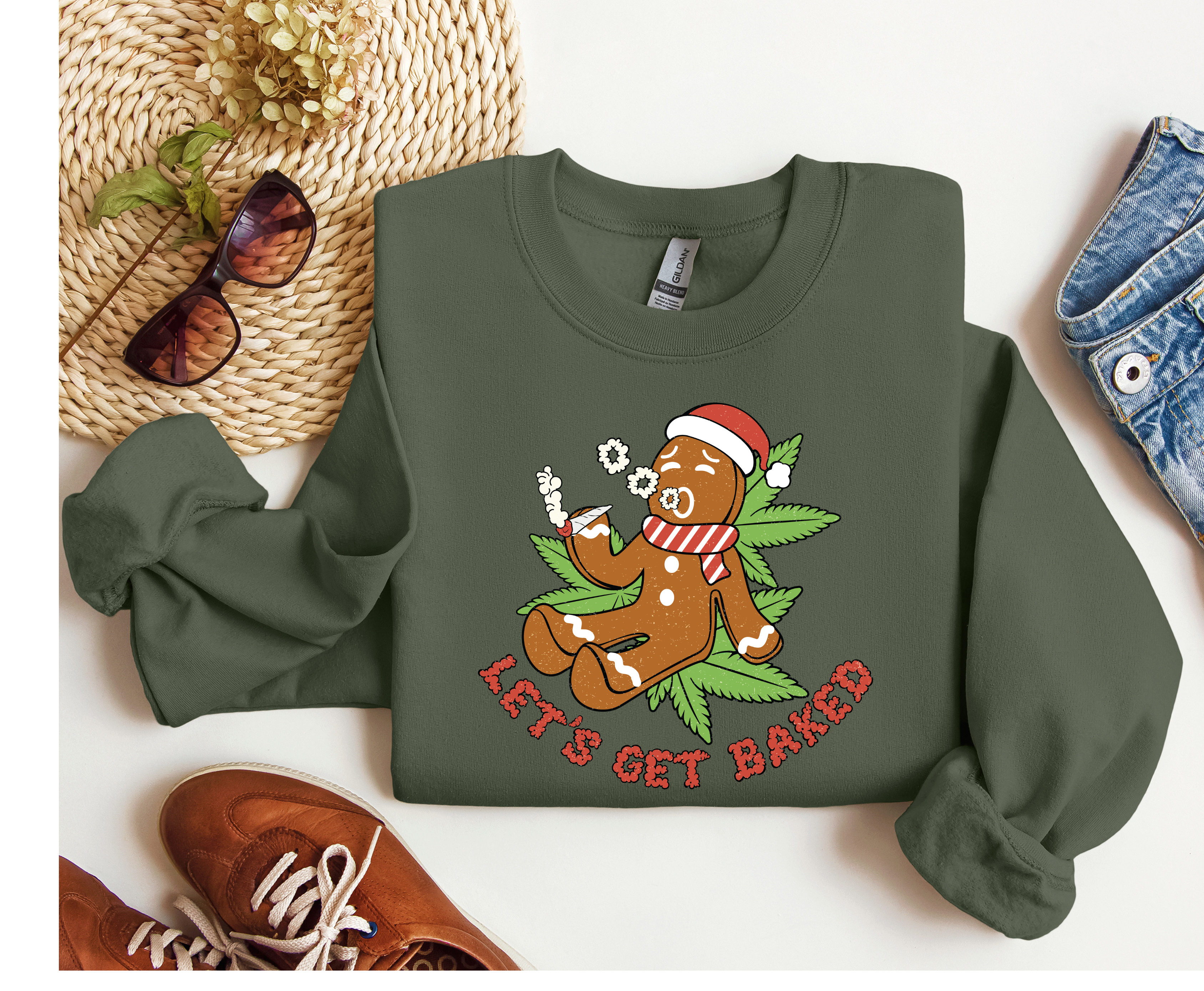 A military green green crewneck sweatshirt laid flat on a white background, styled with sunglasses, a woven hat, brown shoes, and denim jeans. The sweatshirt features a graphic of a gingerbread man wearing a Santa hat and scarf, smoking a spliff and sitting on marijuana leaf, with the text 'LET'S GET BAKED' in red.