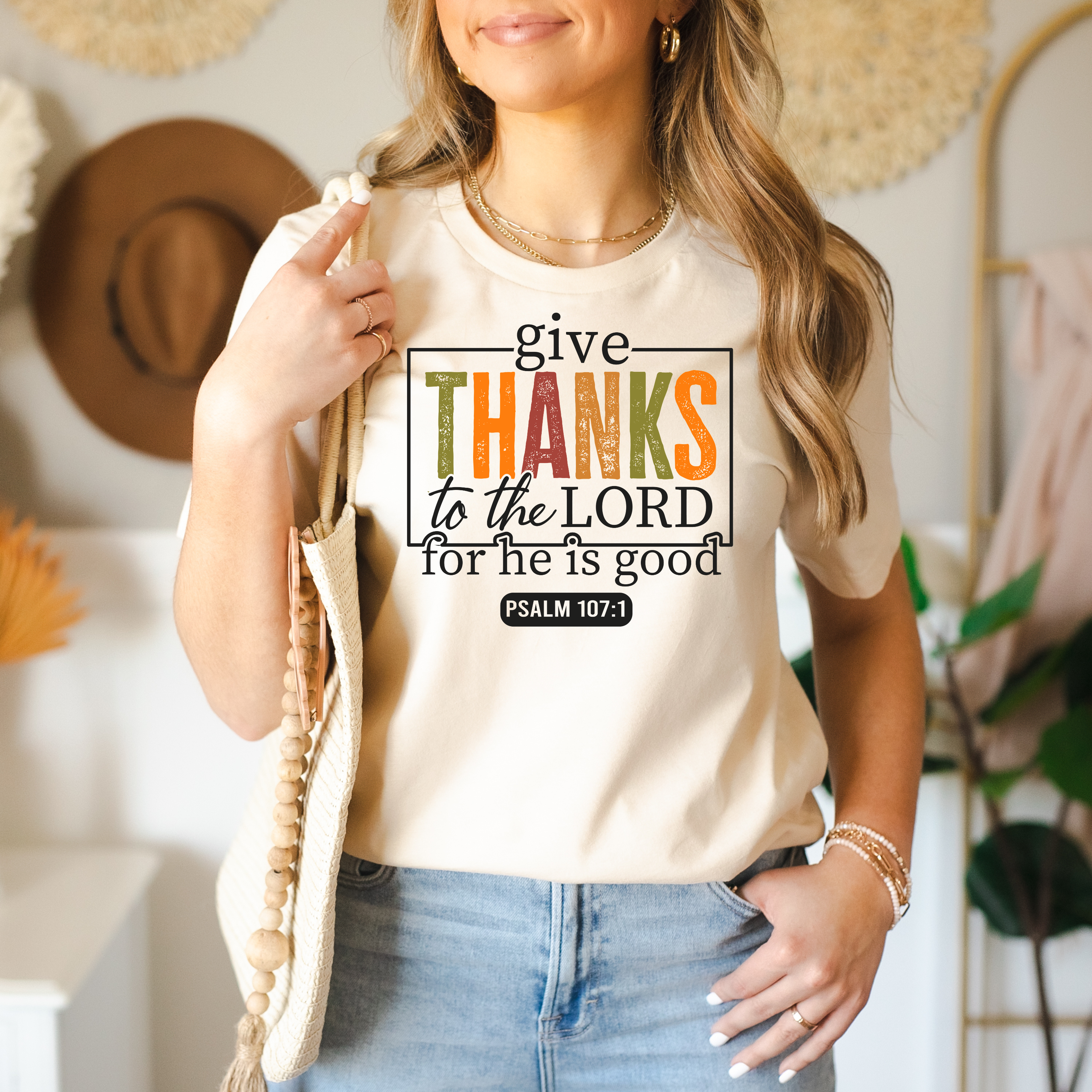A woman wearing a beige graphic t-shirt with a 'give THANKS to the LORD' print, styled with blue jeans and holding a wooden beaded tote bag in a warm, boho-style room.